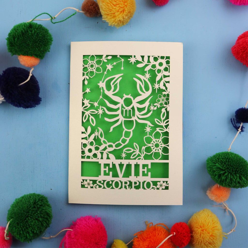 Decorative star sign birthday card with Scorpio symbol and 'EVIE' text on a blue background with colourful pom-poms.