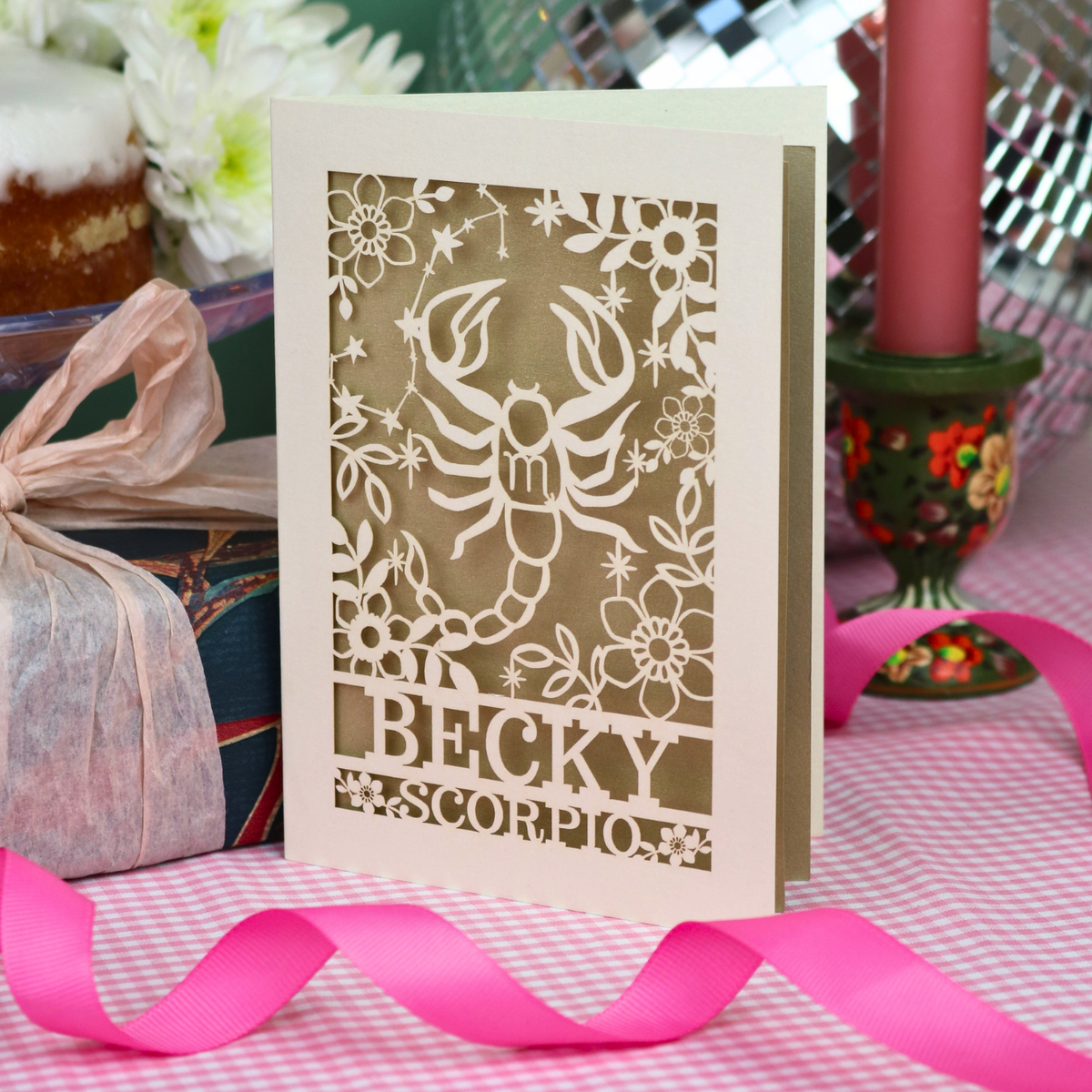 A handmade laser-cut greeting card featuring a cream Scorpio zodiac design, with the name 'BECKY' and 'SCORPIO'. The card is styled on a pink-and-white checked tablecloth with a pink ribbon, surrounded by party decorations.