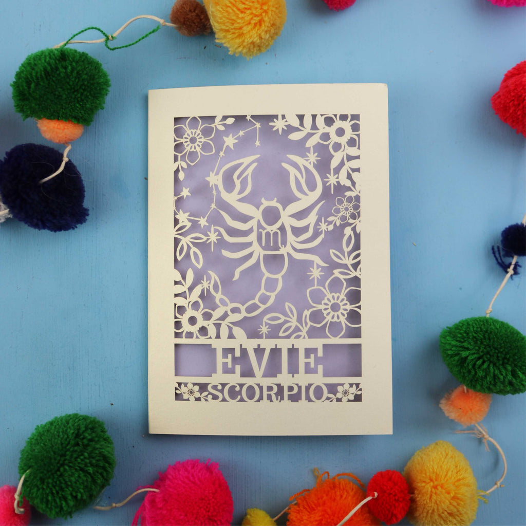 Decorative star sign birthday card with Scorpio symbol and 'EVIE' text on a blue background with colourful pom-poms.