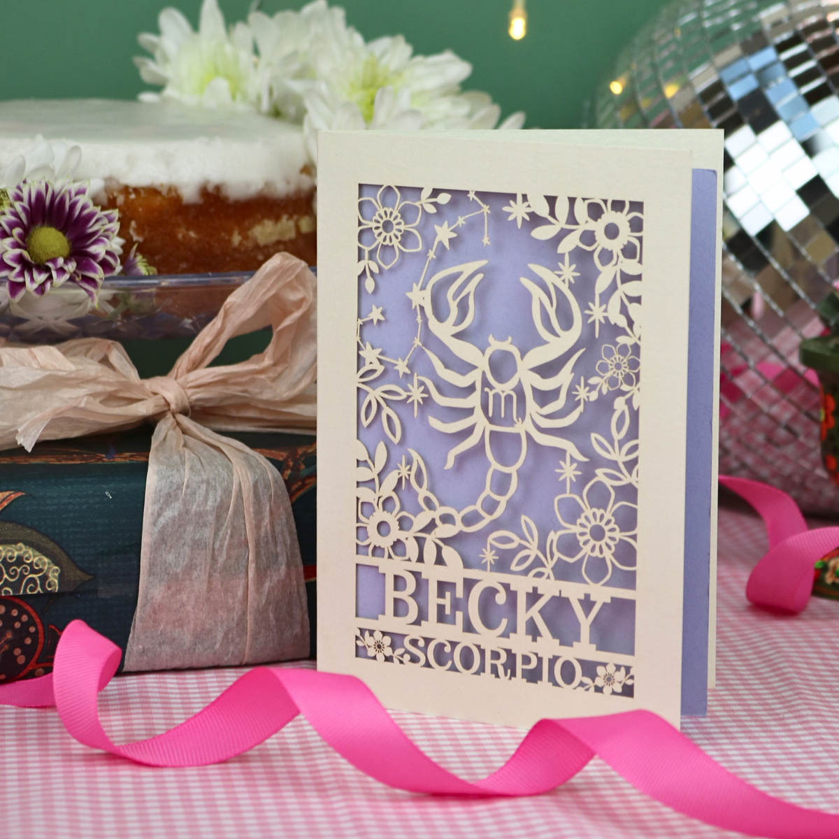 A handmade laser-cut greeting card featuring a cream Scorpio zodiac design over a lilac background, with the name 'BECKY' and 'SCORPIO'. The card is styled on a pink-and-white checked tablecloth with a pink ribbon, surrounded by party decorations.