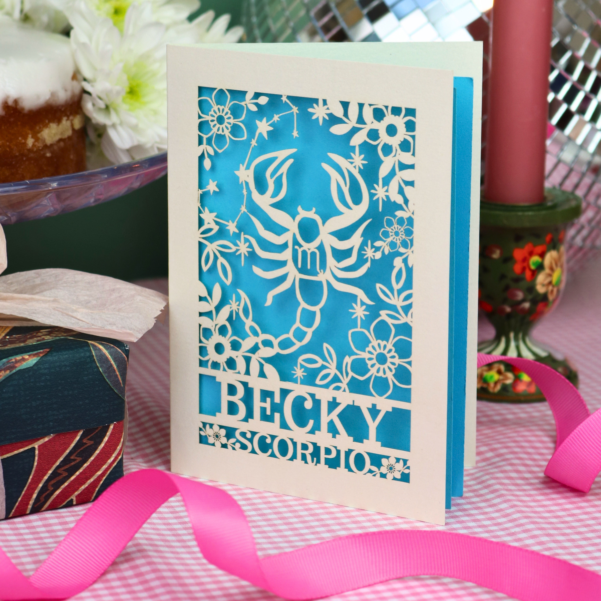 A handmade laser-cut greeting card featuring a cream Scorpio zodiac design over a bright blue background, with the name 'BECKY' and 'SCORPIO'. The card is styled on a pink-and-white checked tablecloth with a pink ribbon, surrounded by party decorations.