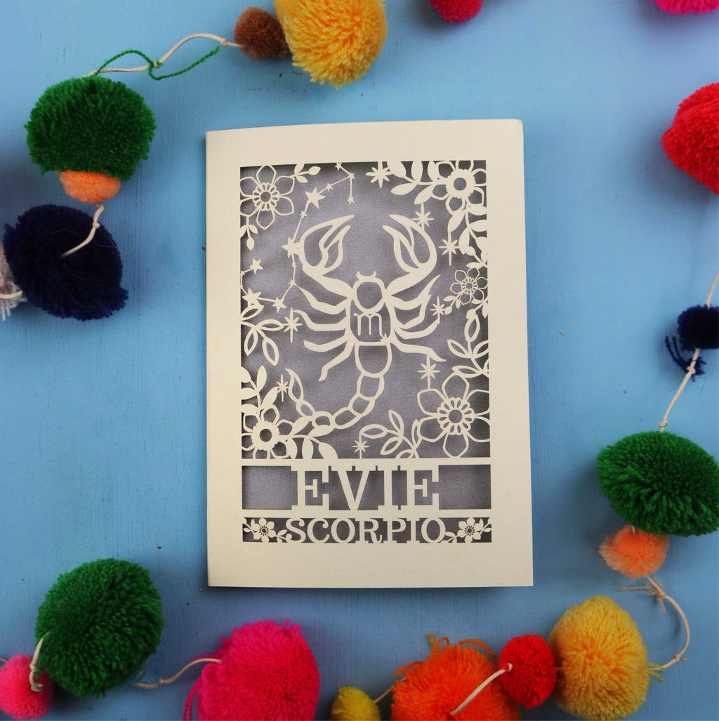 Decorative star sign birthday card with Scorpio symbol and 'EVIE' text on a blue background with colourful pom-poms.