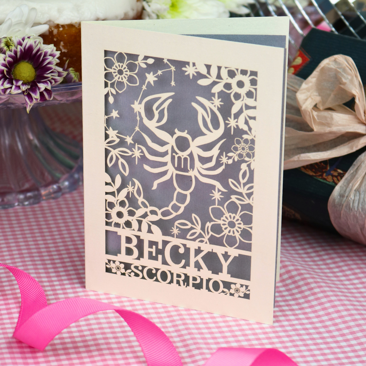 A handmade laser-cut greeting card featuring a cream Scorpio zodiac design over a silver background, with the name 'BECKY' and 'SCORPIO'. The card is styled on a pink-and-white checked tablecloth with a pink ribbon, surrounded by party decorations.