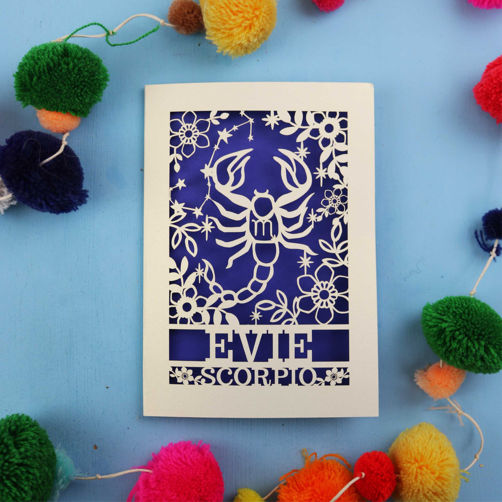 Decorative Scorpio birthday card with a scorpion star sign design and 'Evie Scorpio' text on a blue background with colourful pom-poms.
