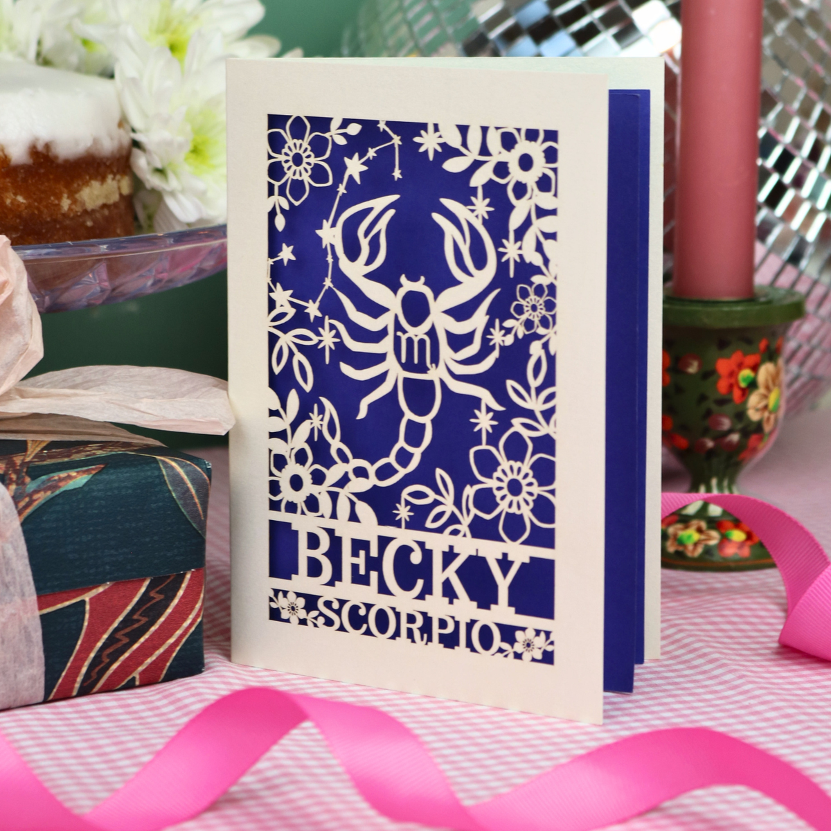 A handmade laser-cut greeting card featuring a cream Scorpio zodiac design over a violet background, with the name 'BECKY' and 'SCORPIO'. The card is styled on a pink-and-white checked tablecloth with a pink ribbon, surrounded by party decorations.