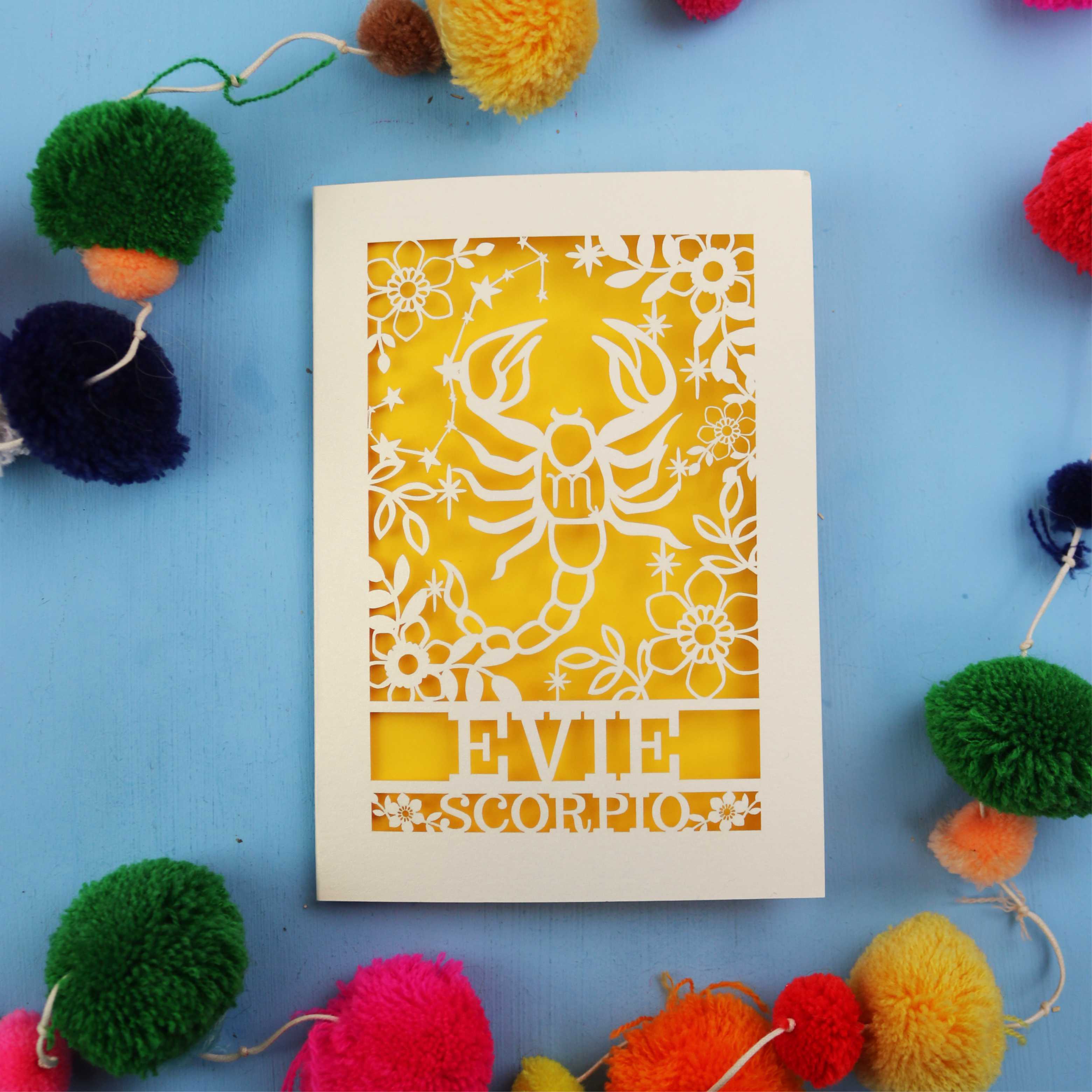 Decorative star sign birthday card with Scorpio symbol and 'EVIE' text on a blue background with colourful pom-poms.