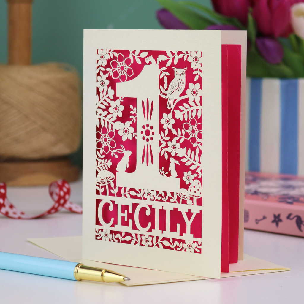 A laser cut 1st birthday card with decorative 1 and woodland design including owl, bunnies, squirrel and hedgehog, personalised with a name.