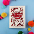 Decorative Valentine's card with 'You are so loved' text on a blue background with colourful pom-poms.