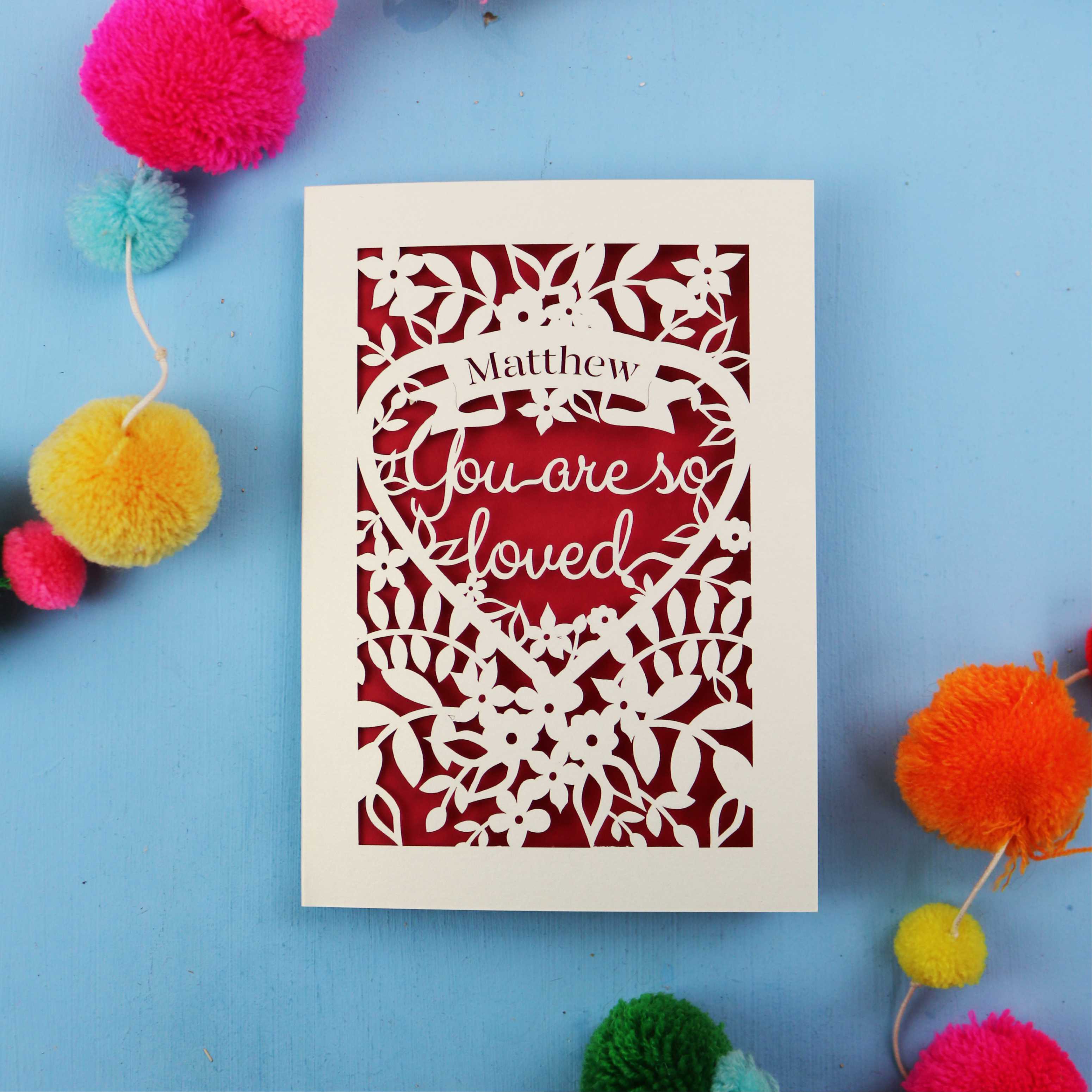Decorative Valentine's card with 'You are so loved' text on a blue background with colourful pom-poms.