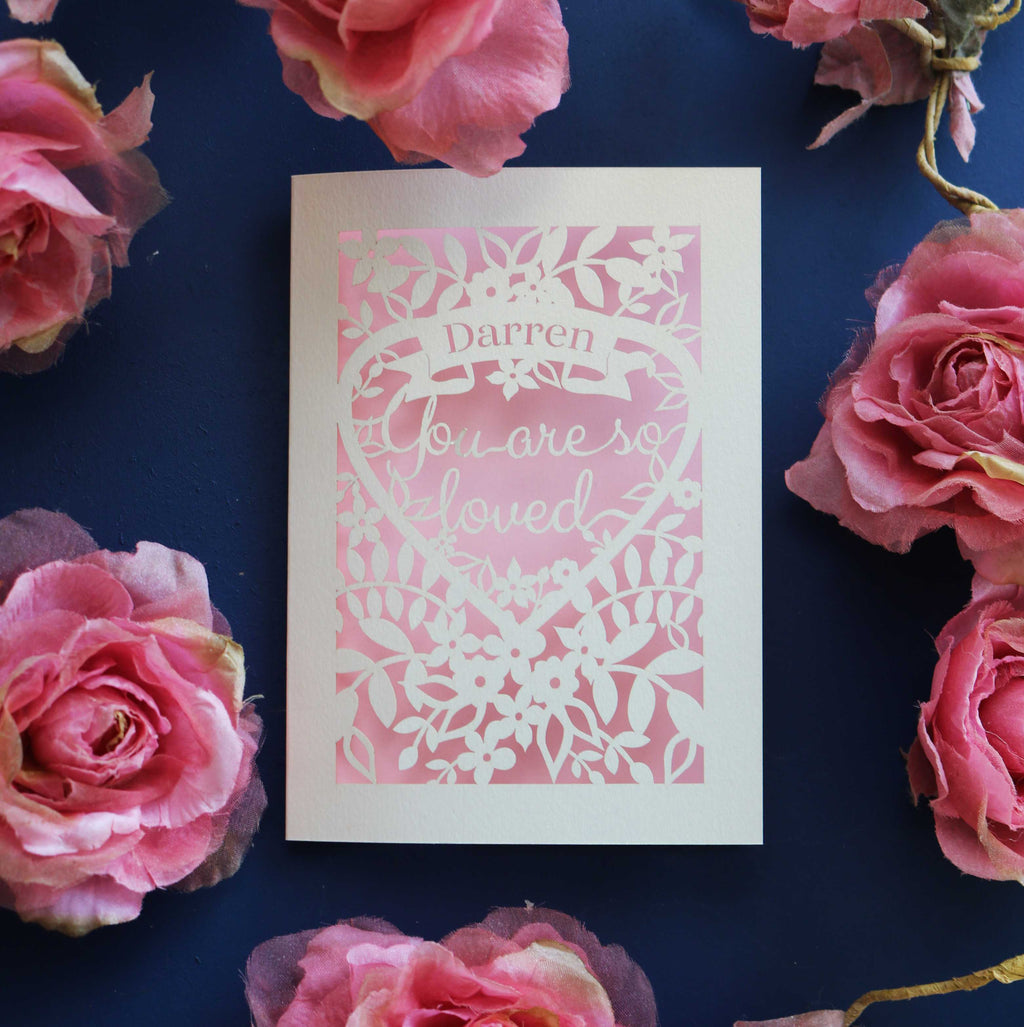 A paper cut Valentine's card personalised with a name. Card says "Name, you are so loved"