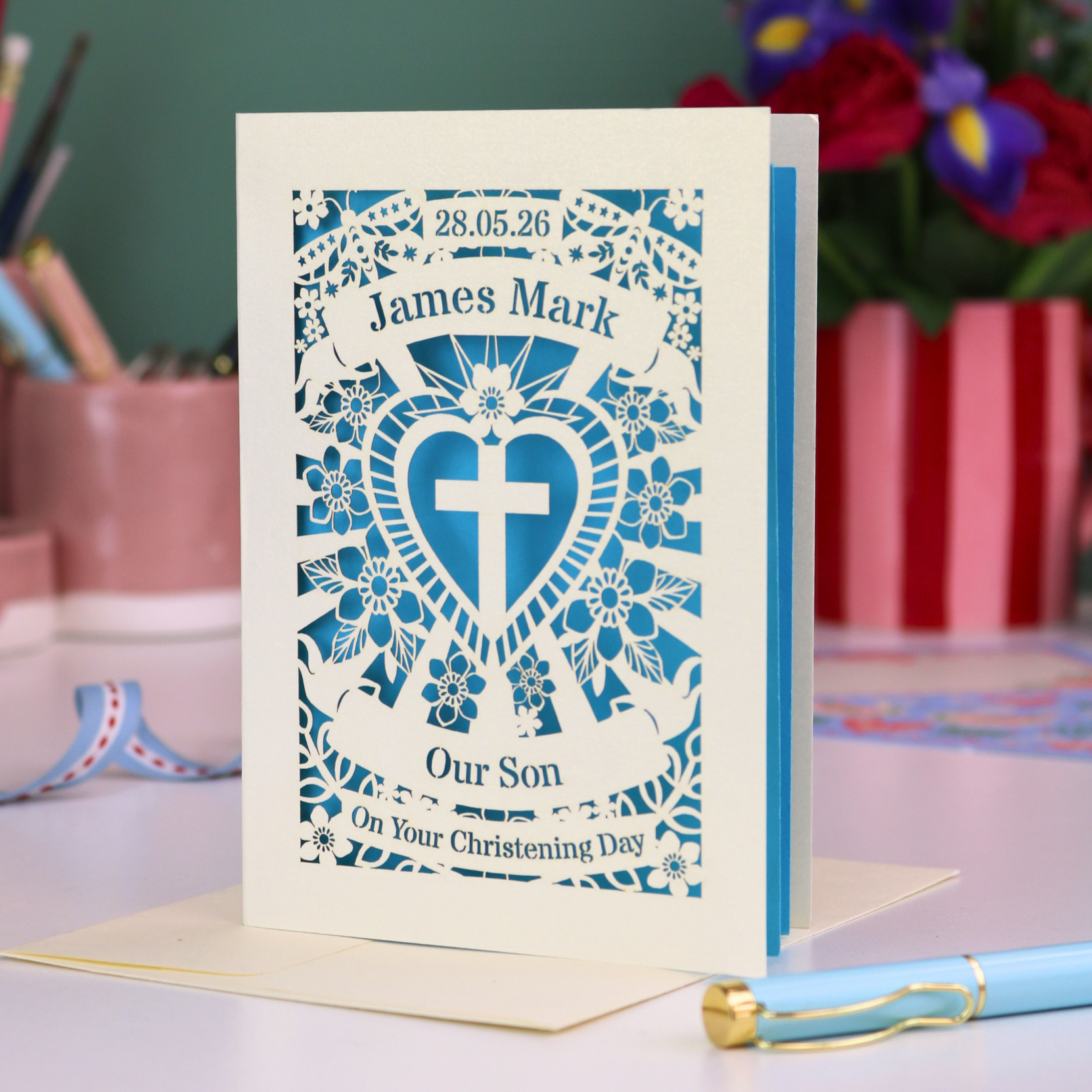 Personalized Son christening card with decorative design and text on a table with flowers in the background.