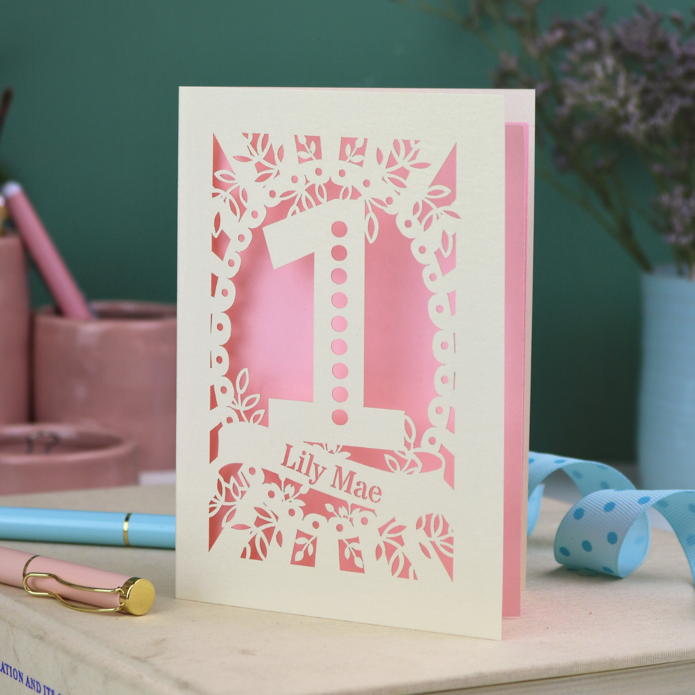 Baby's First Birthday Card with laser cut design and 'Lily Mae' text on a wooden surface with stationery items.