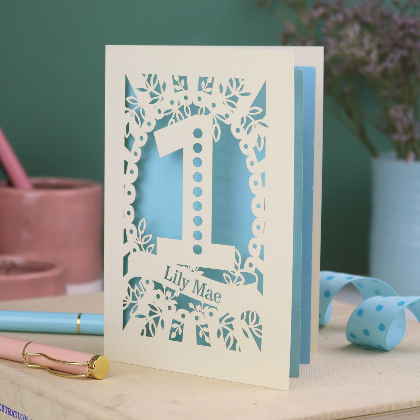 A cream laser cut 1st birthday card with floral border, a spotty number '1', and personalised with the name "Lily Mae" in a stencil font cut out lettering.