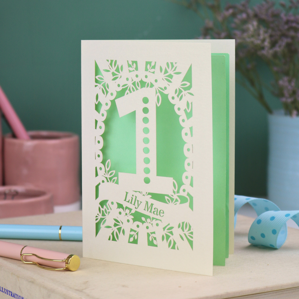 A cream laser cut 1st birthday card with floral border, a spotty number '1', and personalised with the name "Lily Mae" in a stencil font cut out lettering.