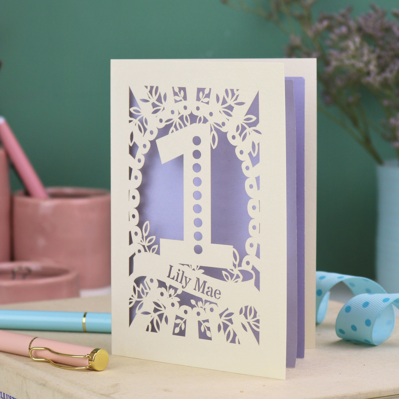 A cream laser cut 1st birthday card with floral border, a spotty number '1', and personalised with the name "Lily Mae" in a stencil font cut out lettering.
