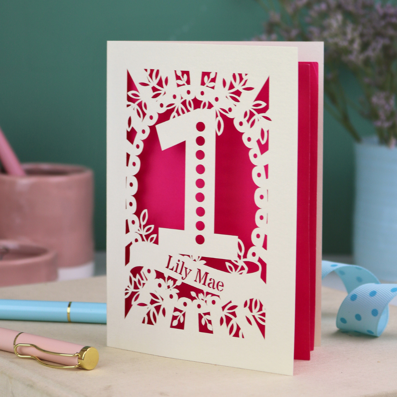 A cream laser cut first birthday card with floral border, a spotty number '1', and personalised with the name "Lily Mae" in a stencil font cut out lettering.
