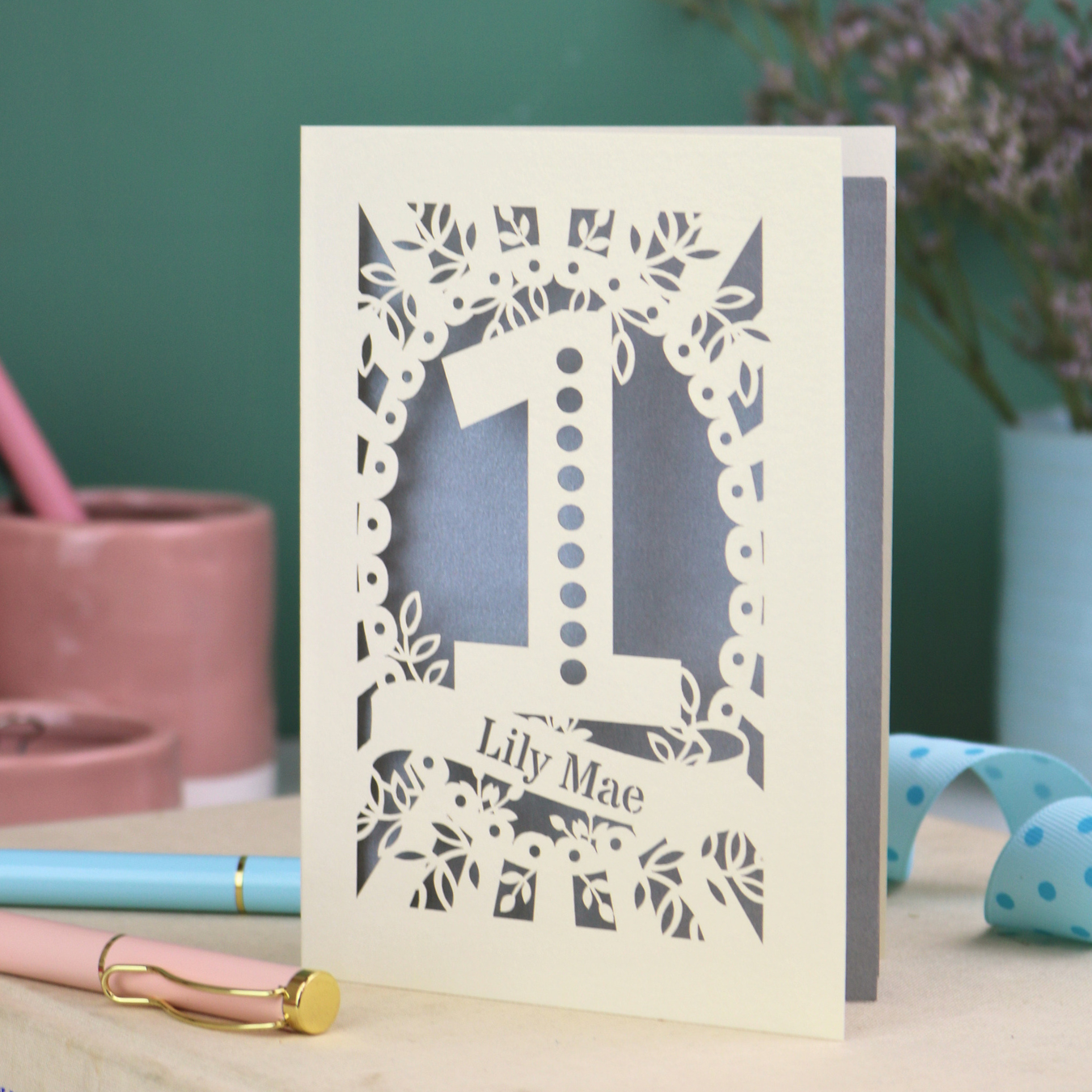A cream laser cut 1st birthday card with floral border, a spotty number '1', and personalised with the name "Lily Mae" in a stencil font cut out lettering.