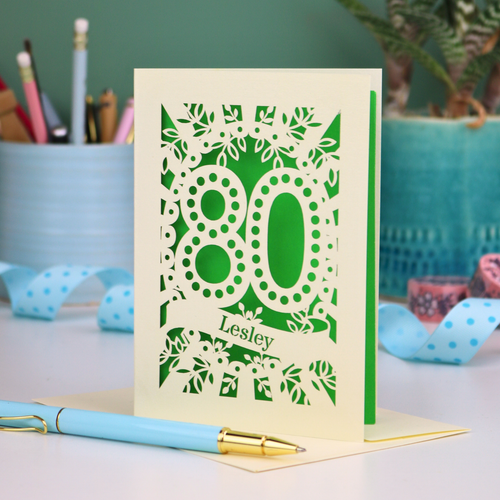 Laser cut cream and Green personalised 80th birthday card with decorative patterns on a desk with stationery items.