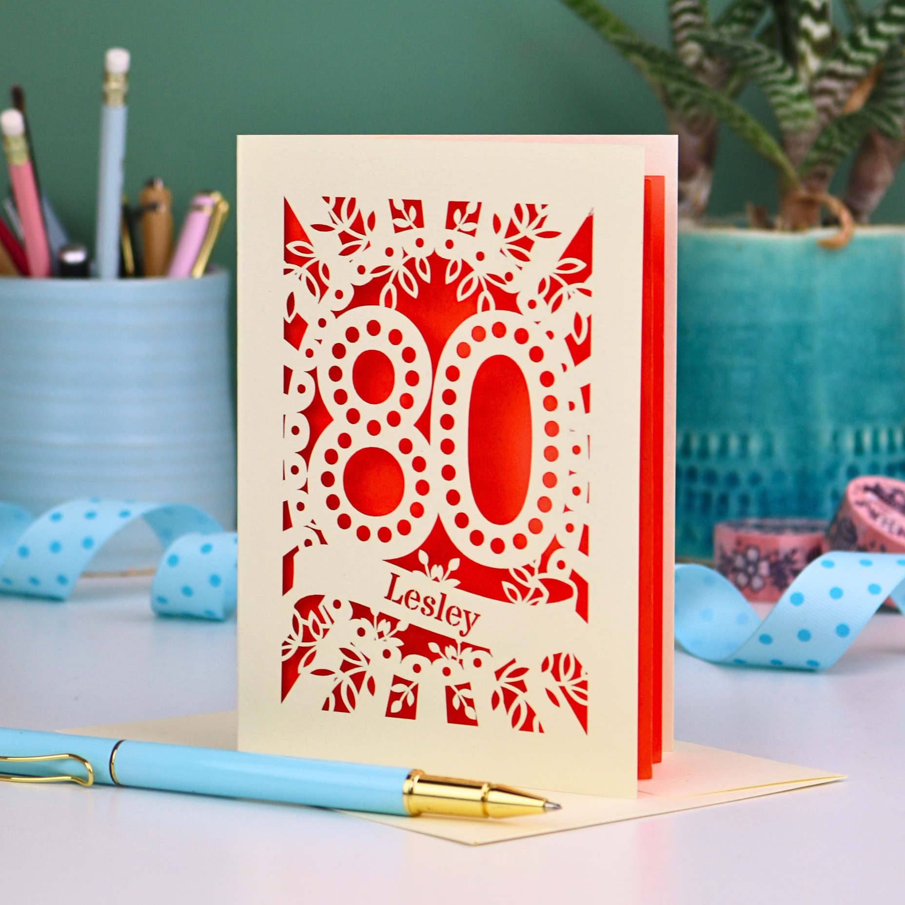 Laser cut cream and orange personalised 80th birthday card with decorative patterns on a desk with stationery items.