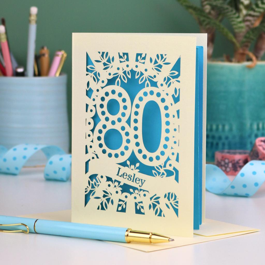 Laser cut cream and blue personalised 80th birthday card with decorative patterns on a desk with stationery items.