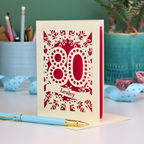 Laser cut cream and pink personalised 80th birthday card with decorative patterns on a desk with stationery items.