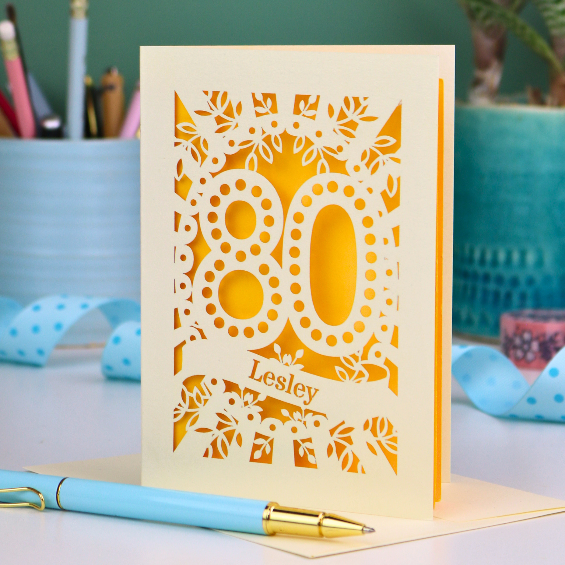 Laser cut cream and yellow personalised 80th birthday card with decorative patterns on a desk with stationery items.