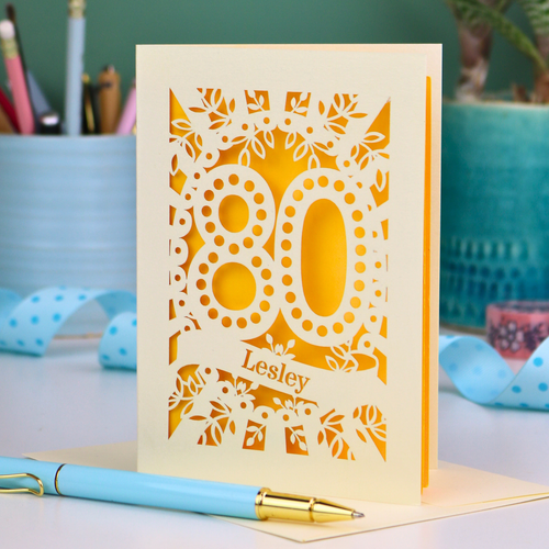 Laser cut cream and yellow personalised 80th birthday card with decorative patterns on a desk with stationery items.