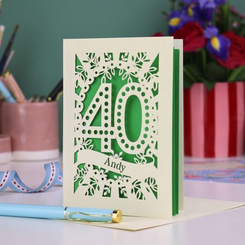 Laser-cut 40th birthday card with number 40 and a stencil cut out name, on a desk with stationery items and flowers.