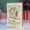 Laser-cut 40th birthday card with number 40 and a stencil cut out name, on a desk with stationery items and flowers.