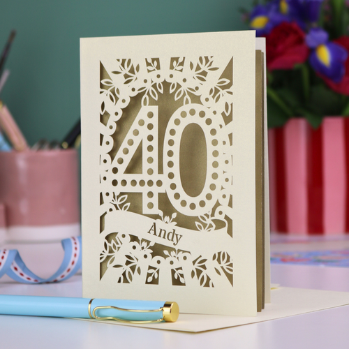 Laser-cut 40th birthday card with number 40 and a stencil cut out name, on a desk with stationery items and flowers.
