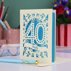 Laser-cut 40th birthday card with number 40 and a stencil cut out name, on a desk with stationery items and flowers.