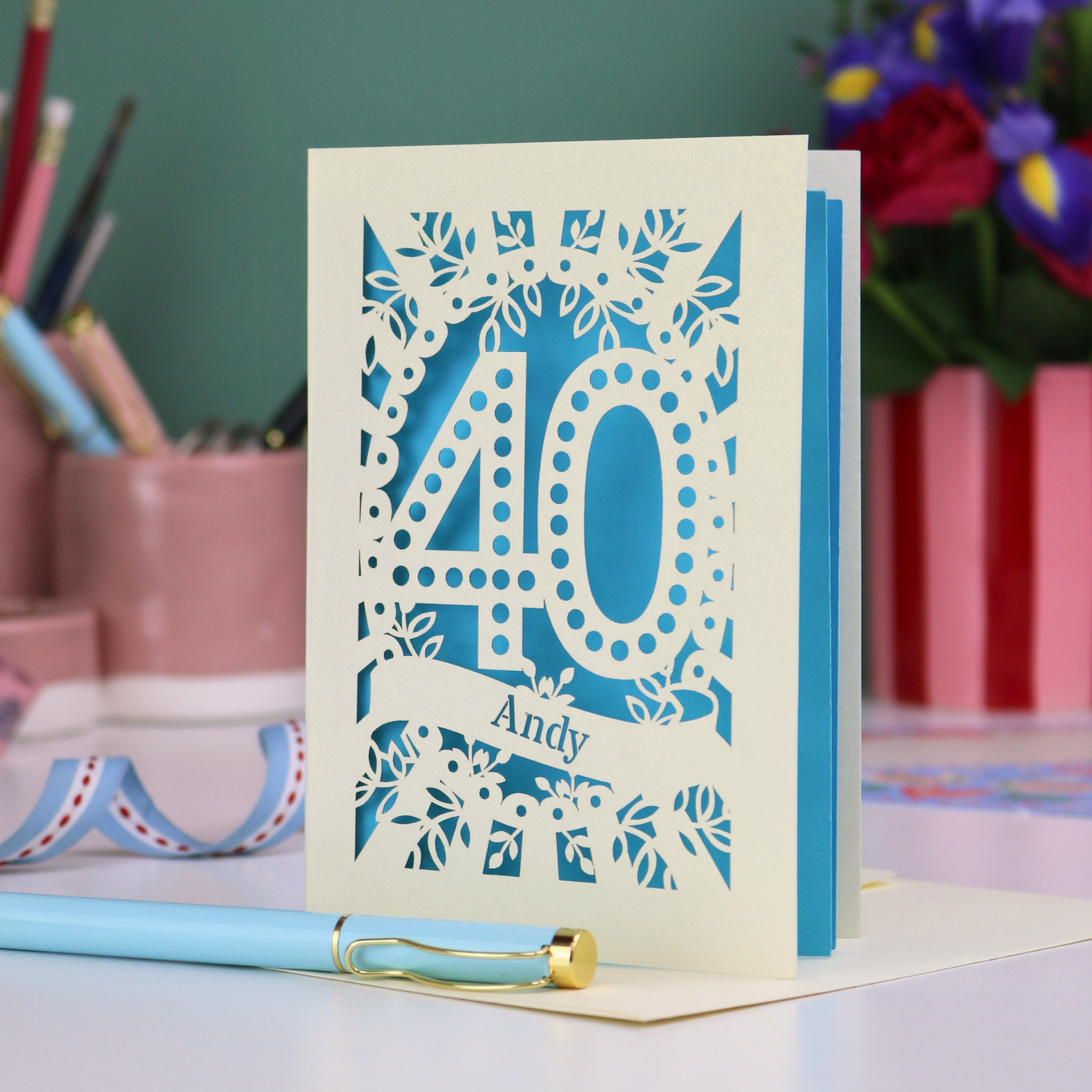 Laser-cut 40th birthday card with number 40 and a stencil cut out name, on a desk with stationery items and flowers.