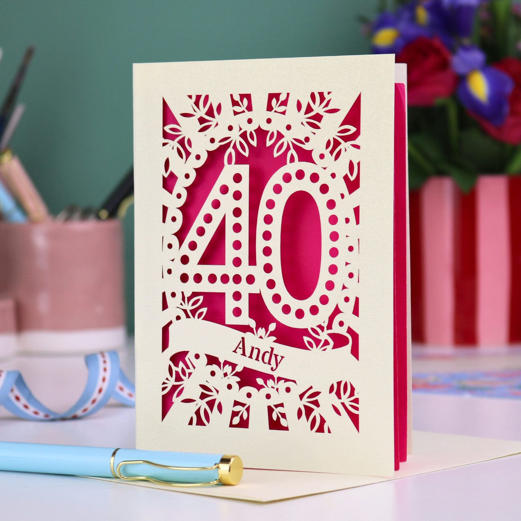 Laser-cut 40th birthday card with number 40 and a stencil cut out name, on a desk with stationery items and flowers.