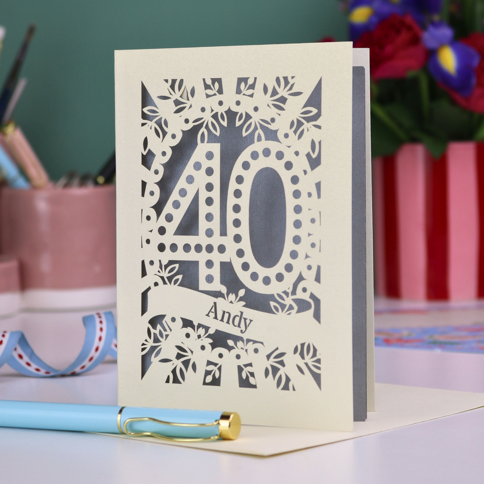 Laser-cut 40th birthday card with number 40 and a stencil cut out name, on a desk with stationery items and flowers.