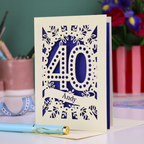 Laser-cut 40th birthday card with number 40 and a stencil cut out name, on a desk with stationery items and flowers.