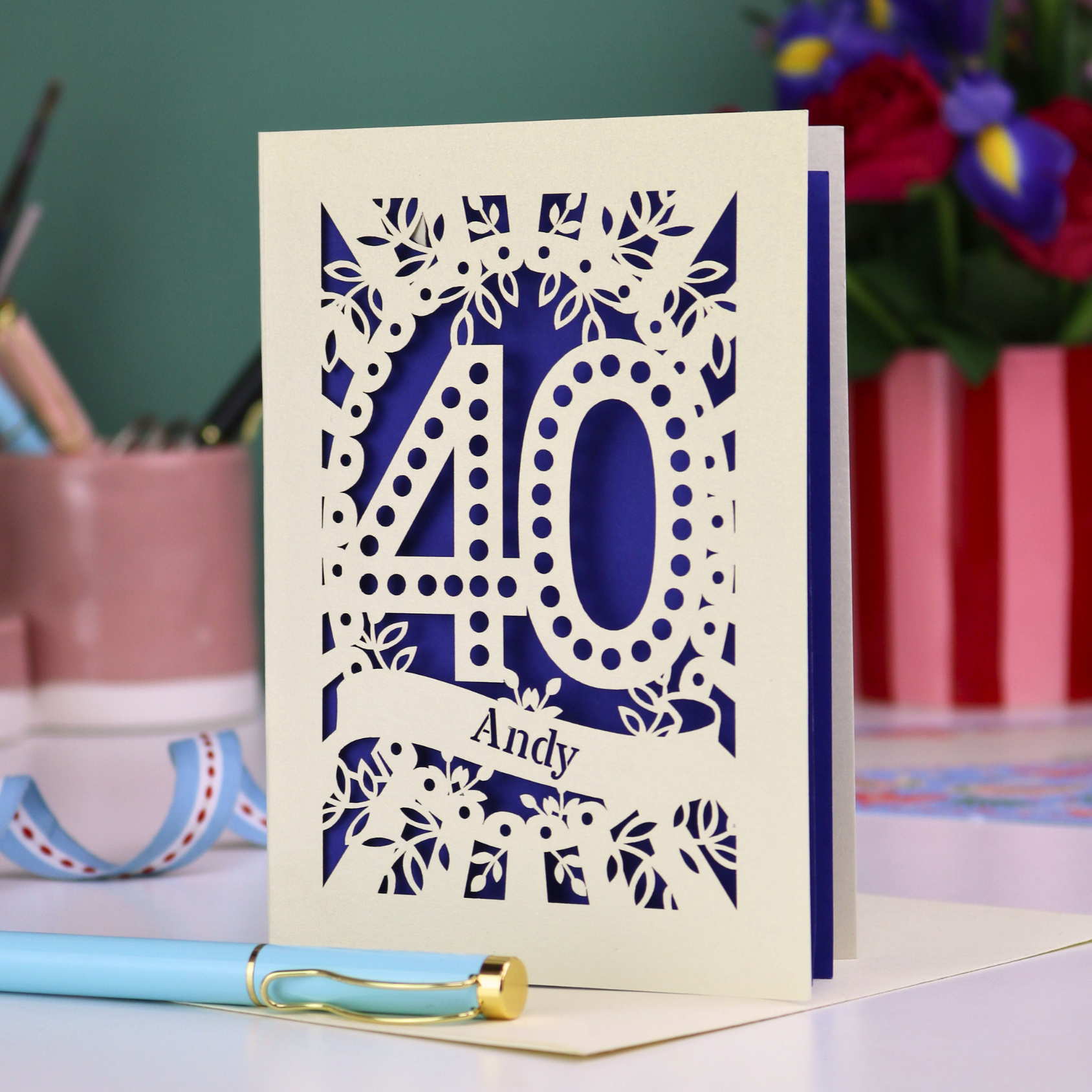 Laser-cut 40th birthday card with number 40 and a stencil cut out name, on a desk with stationery items and flowers.