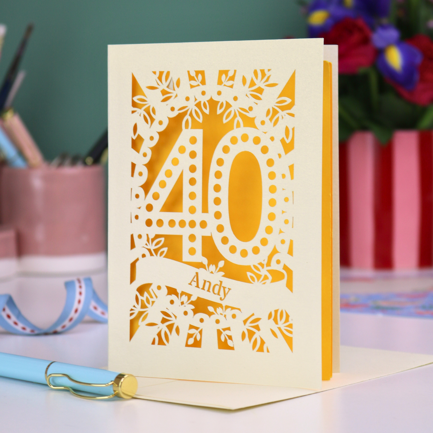Laser-cut 40th birthday card with number 40 and a stencil cut out name, on a desk with stationery items and flowers.