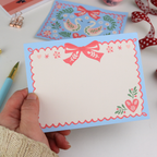 Hand holding a decorative notecard with a pink bow and floral design on a white surface.