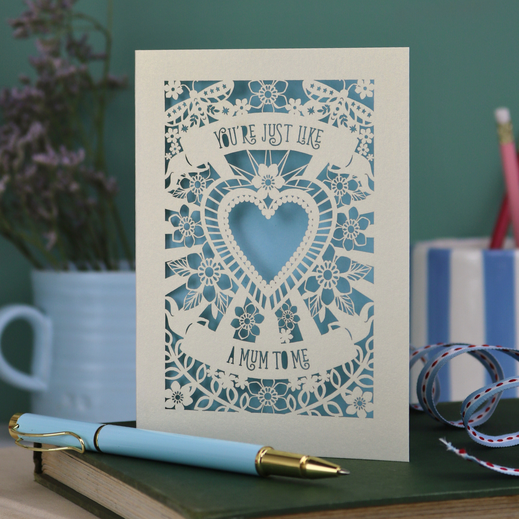 Decorative Step-mum Mother's Day card with floral design and text 'You're just like a mum to me' on a green surface with a pen and mug in the background.