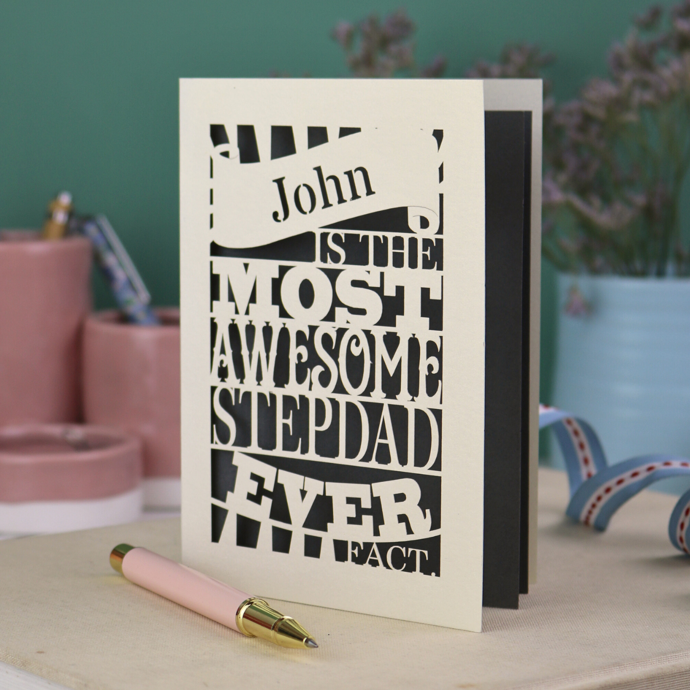 Paper cut Stepdad Father's day Card with personalized text, "John is the most awesome stepdad ever. Fact." on a table with decorative items