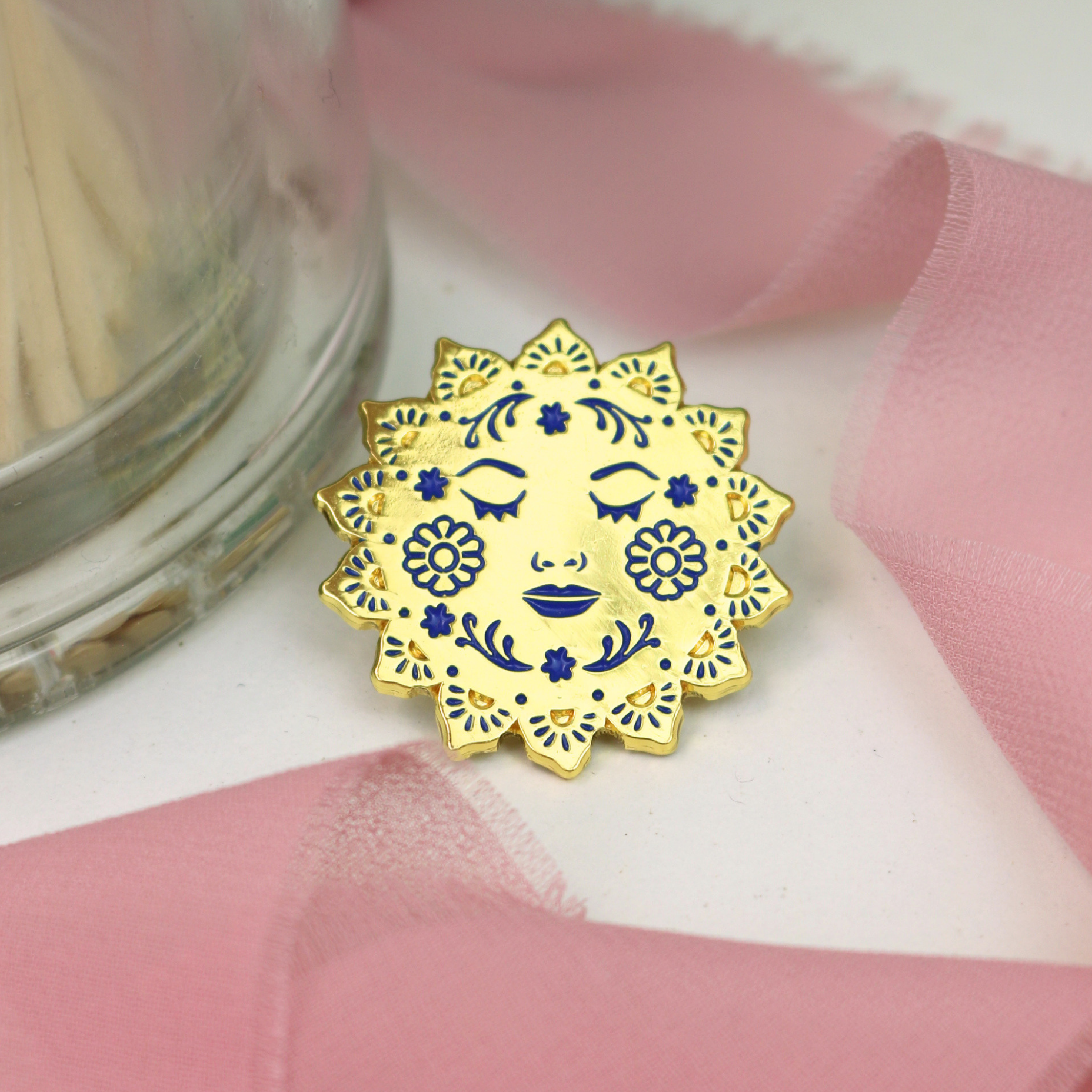 Decorative sun-shaped enamel lapel pin with face design on a textured fabric background
