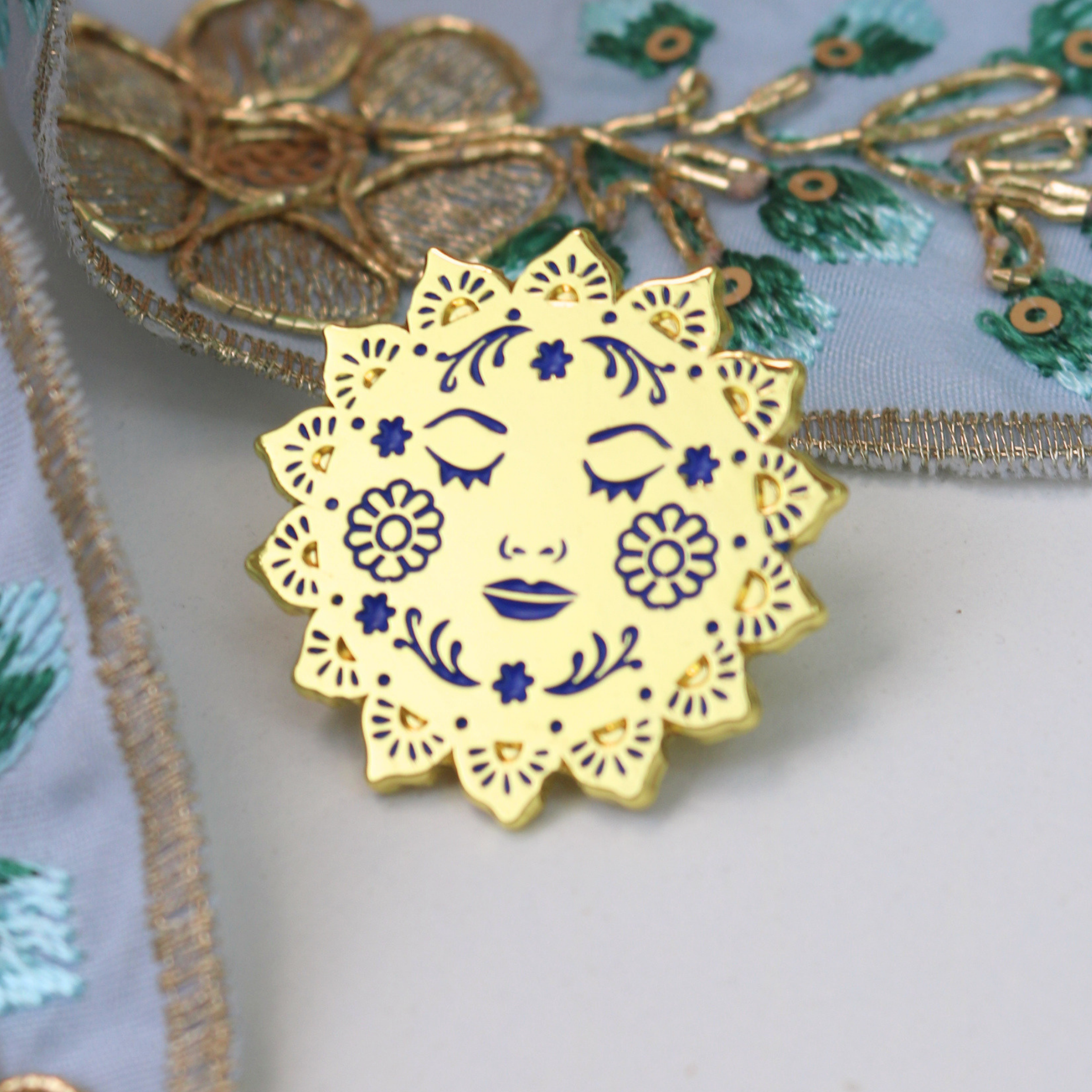 Decorative sun-shaped enamel lapel pin with face design on a textured fabric background