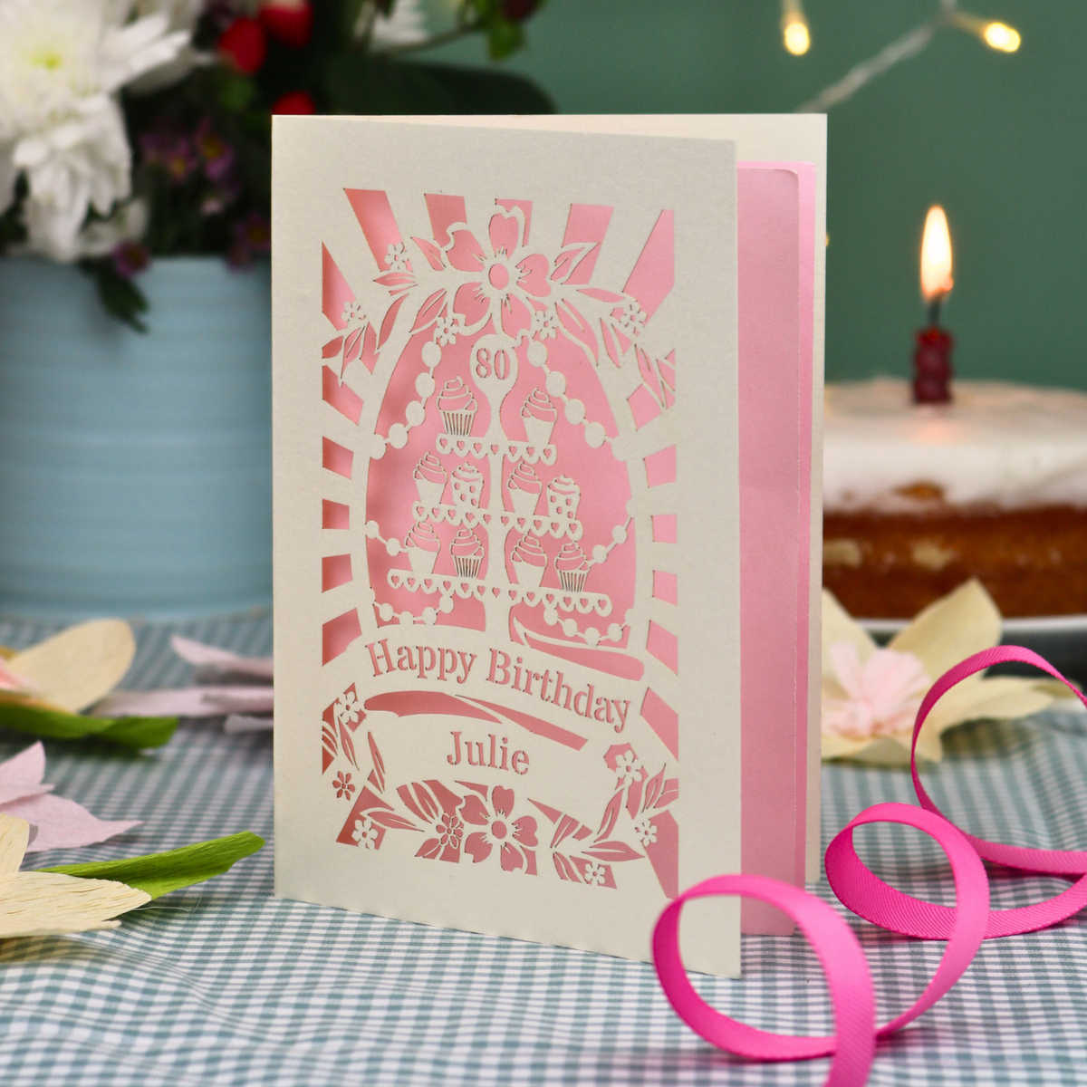 A personalised laser-cut 80th birthday card with a cream exterior and pastel pink interior, featuring a cupcake stand design and the text 'Happy Birthday Julie'. The card is displayed on a blue-and-white checked tablecloth with a pink ribbon, with a blurred birthday cake and candle in the background.