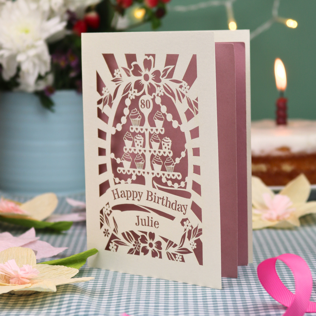 A personalised cut out 80th birthday card with a cream exterior and dusky pink interior, featuring a cupcake stand design and the text 'Happy Birthday Julie'. The card is displayed on a blue-and-white checked tablecloth with a pink ribbon, with a blurred birthday cake and candle in the background.
