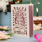 A personalised cut out 80th birthday card with a cream exterior and dusky pink interior, featuring a cupcake stand design and the text 'Happy Birthday Julie'. The card is displayed on a blue-and-white checked tablecloth with a pink ribbon, with a blurred birthday cake and candle in the background.