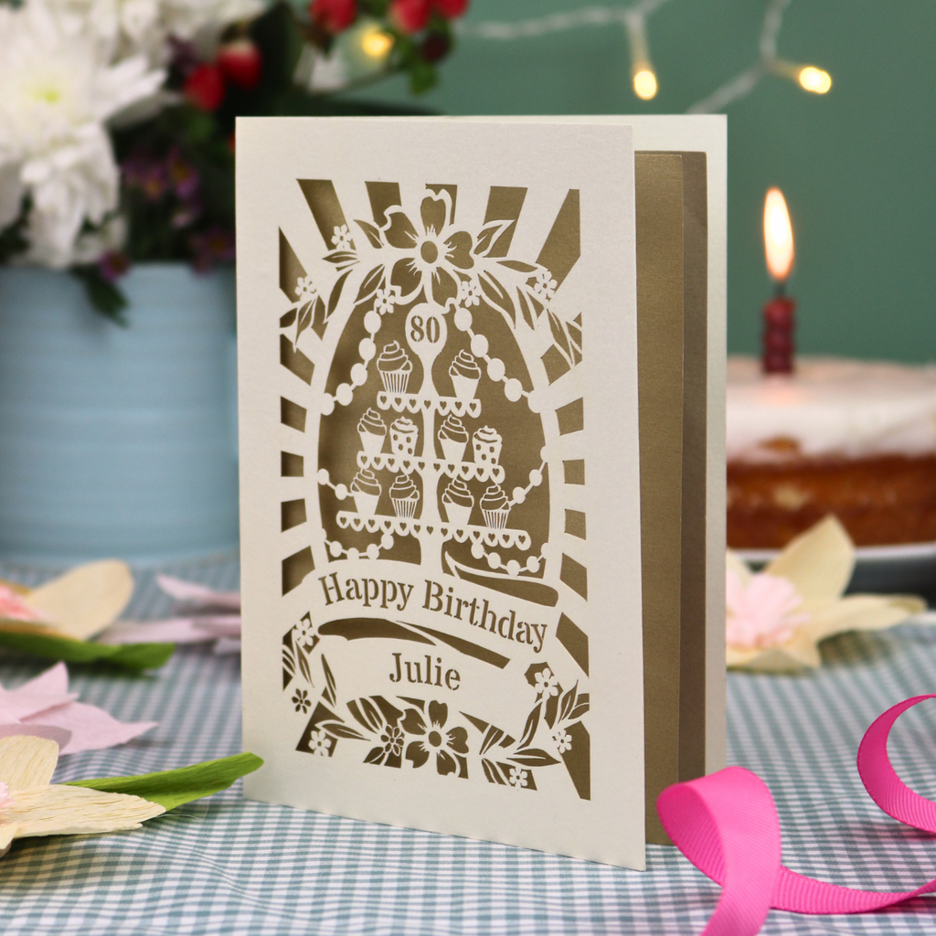 A personalised laser-cut 80th birthday card with a cream exterior and gold leaf interior, featuring a tea party cupcake stand design and the text 'Happy Birthday Julie'. The card is displayed on a blue-and-white checked tablecloth with a pink ribbon, with a blurred birthday cake and candle in the background.