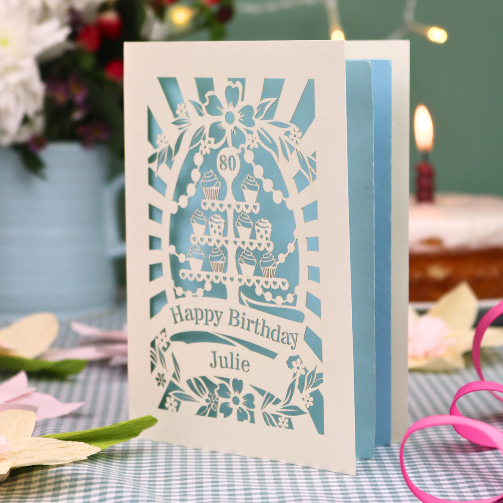 A personalised laser-cut 80th birthday card with a cream exterior and light blue interior, featuring a cupcake stand design and the text 'Happy Birthday Julie'. The card is displayed on a blue-and-white checked tablecloth with a pink ribbon, with a blurred birthday cake and candle in the background.