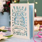 A personalised laser-cut 80th birthday card with a cream exterior and light blue interior, featuring a cupcake stand design and the text 'Happy Birthday Julie'. The card is displayed on a blue-and-white checked tablecloth with a pink ribbon, with a blurred birthday cake and candle in the background.