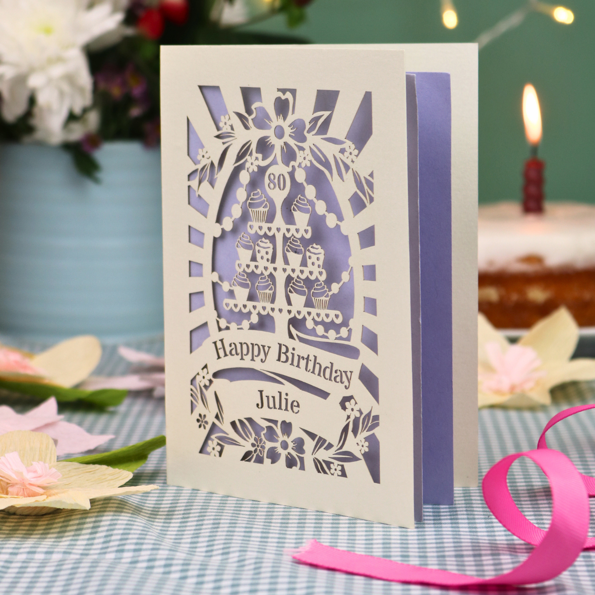A personalised laser-cut 80th birthday card with a cream exterior and lilac interior, featuring a cupcake stand design and the text 'Happy Birthday Julie'. The card is displayed on a blue-and-white checked tablecloth with a pink ribbon, with a blurred birthday cake and candle in the background.