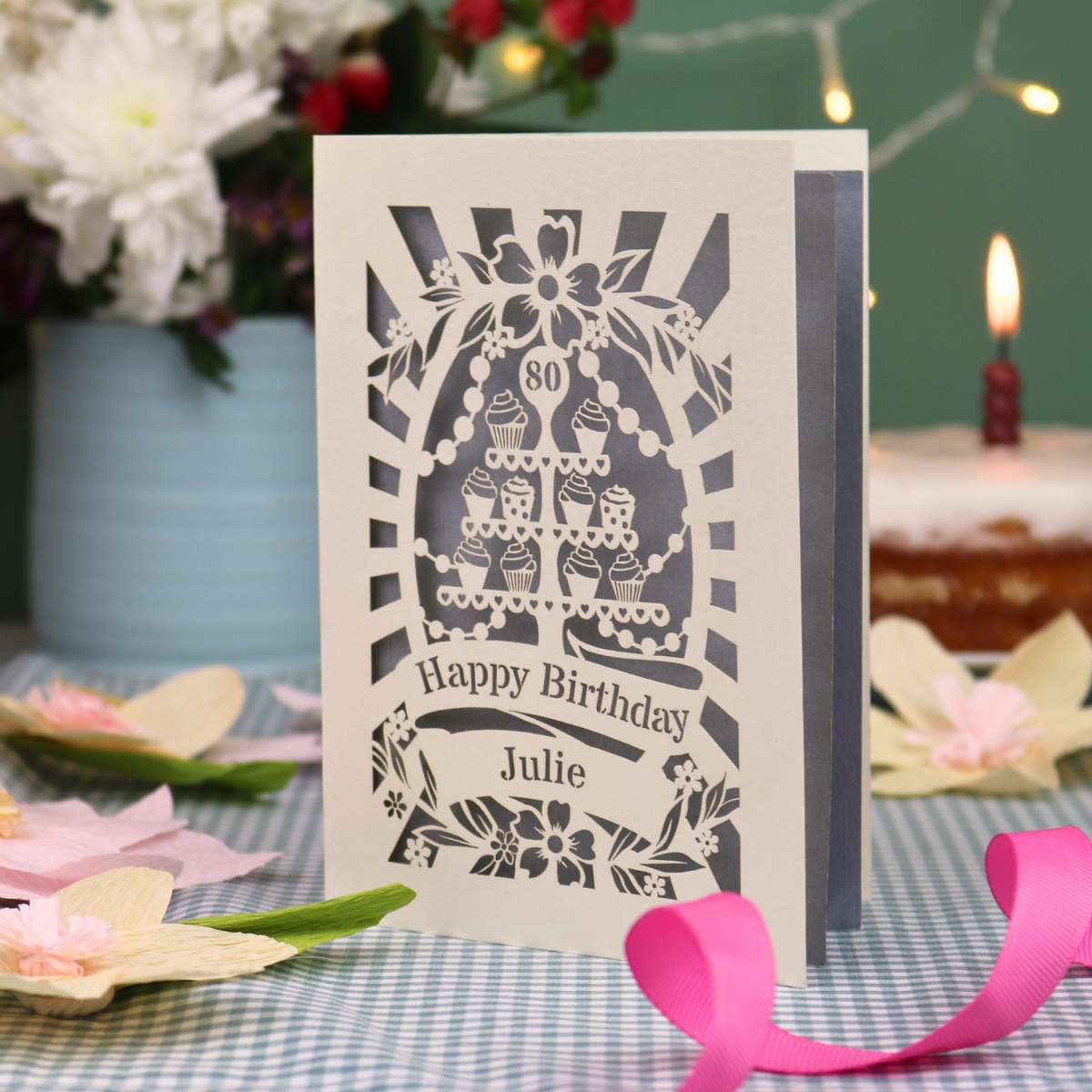 A personalised laser-cut 80th birthday card with a cream exterior and silver interior, featuring a cupcake stand garden party design and the text 'Happy Birthday Julie'. The card is displayed on a blue-and-white checked tablecloth with a pink ribbon, with a blurred birthday cake and candle in the background.